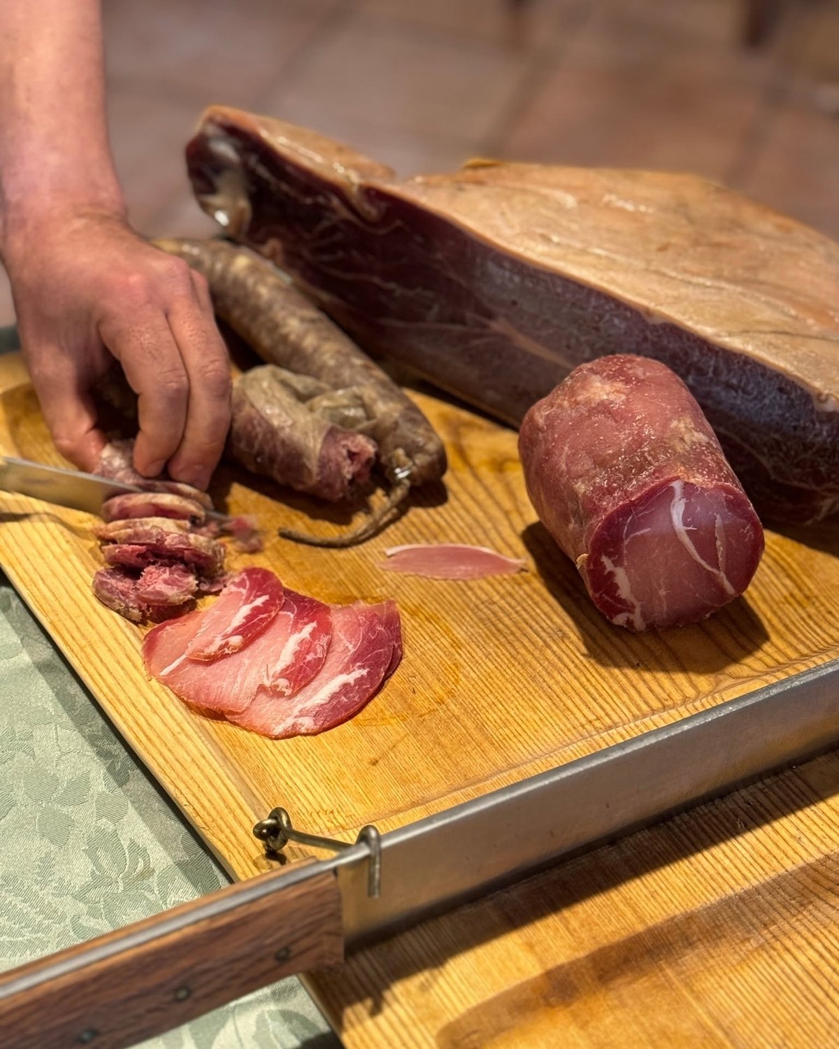 Mano che affetta salsiccia stagionata con il coltello accanto a prosciutto e lonza su un tagliere di legno chiaro