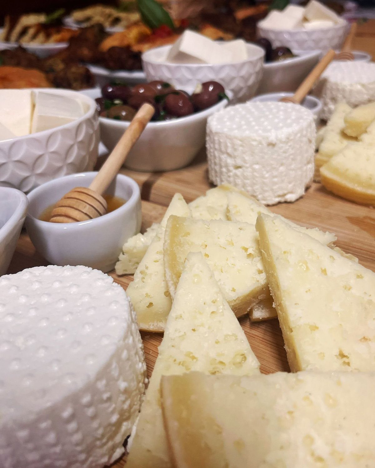 Tagliere di legno con forme di ricotta, spicchi di formaggio stagionato, olive scure e ciotolina di miele con mestolino